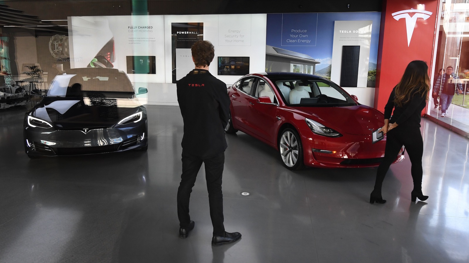 Two Tesla employees stand with their backs to the camera in a Tesla showroom with two Model S sedans.