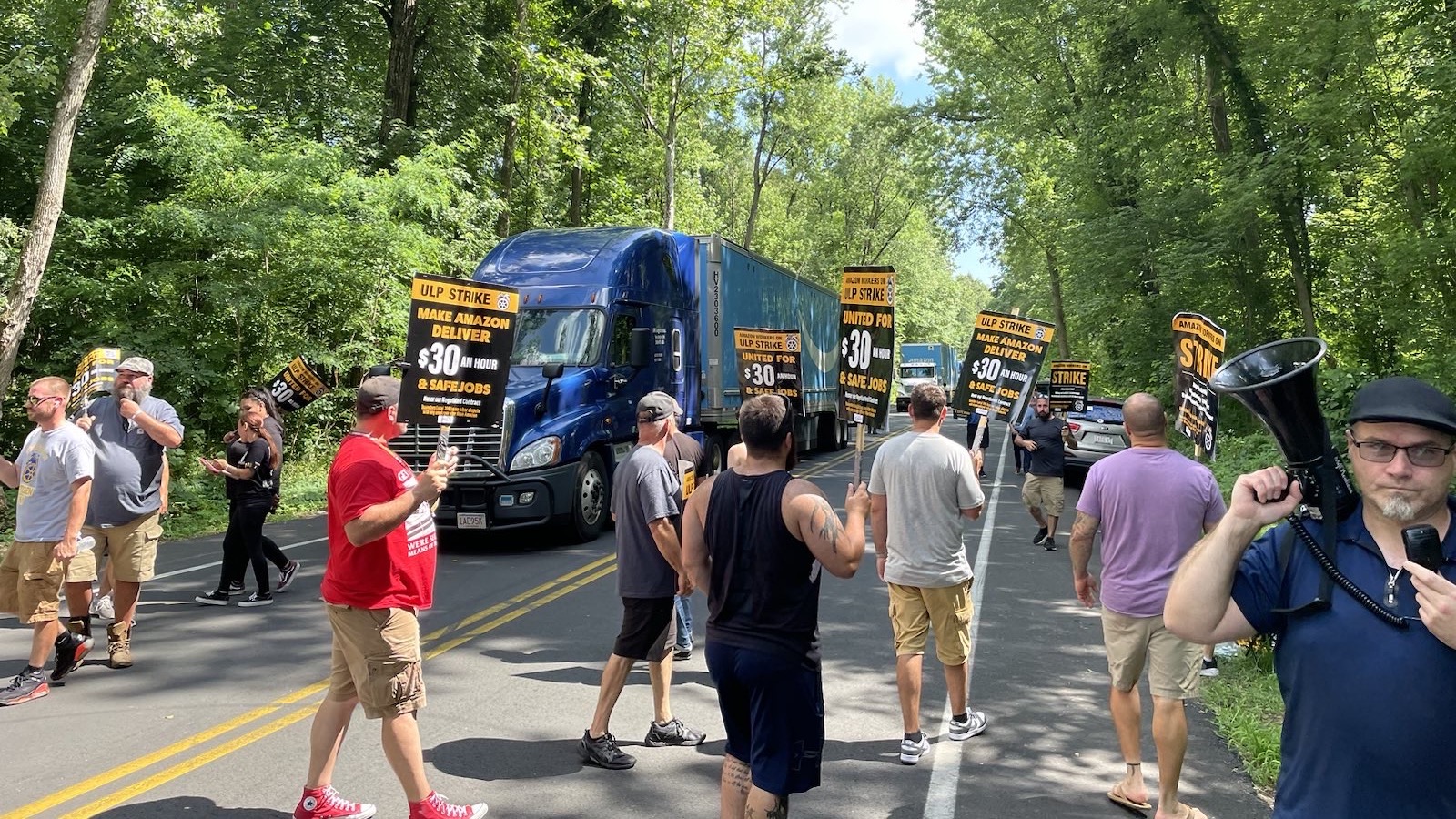 crowd of drivers marching on a road lined with trees in connecticut, holding signs saying make amazon deliver $30 an hour and safe jobs and halting amazon branded semi trucks