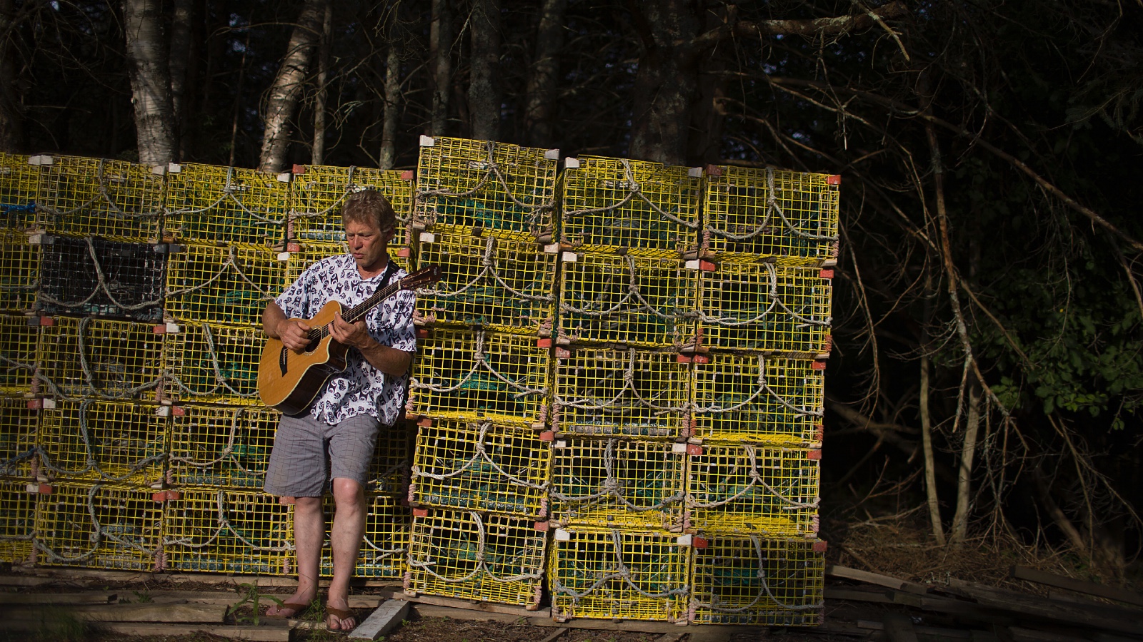 A Maine lobsterman plays guitar in front of rows of stacked lobster traps.