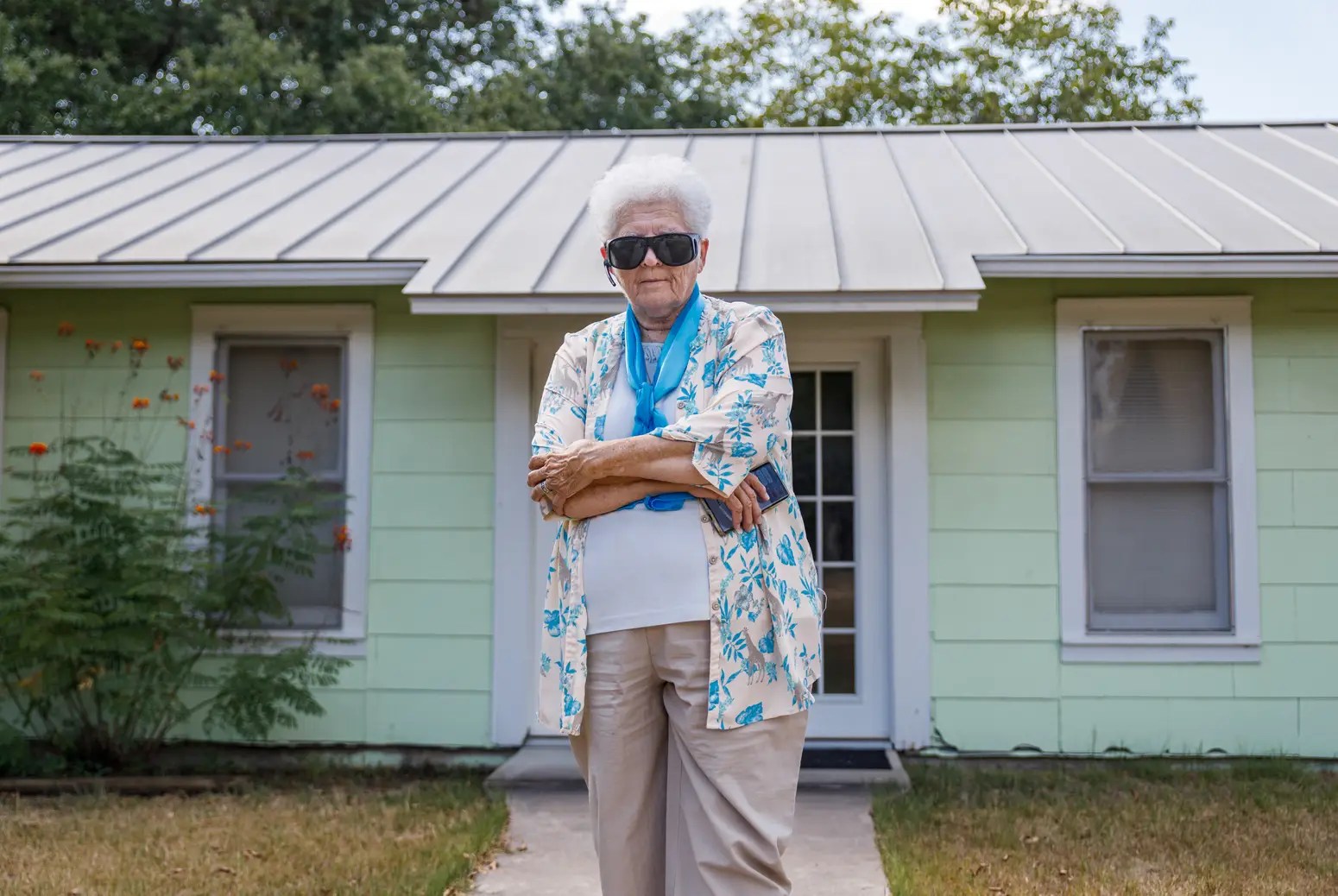 An older woman with white hair, slacks, and a colorful shirt and blue scarf and black sunglasses stands defiantly with her arms crossed in front of a green house.