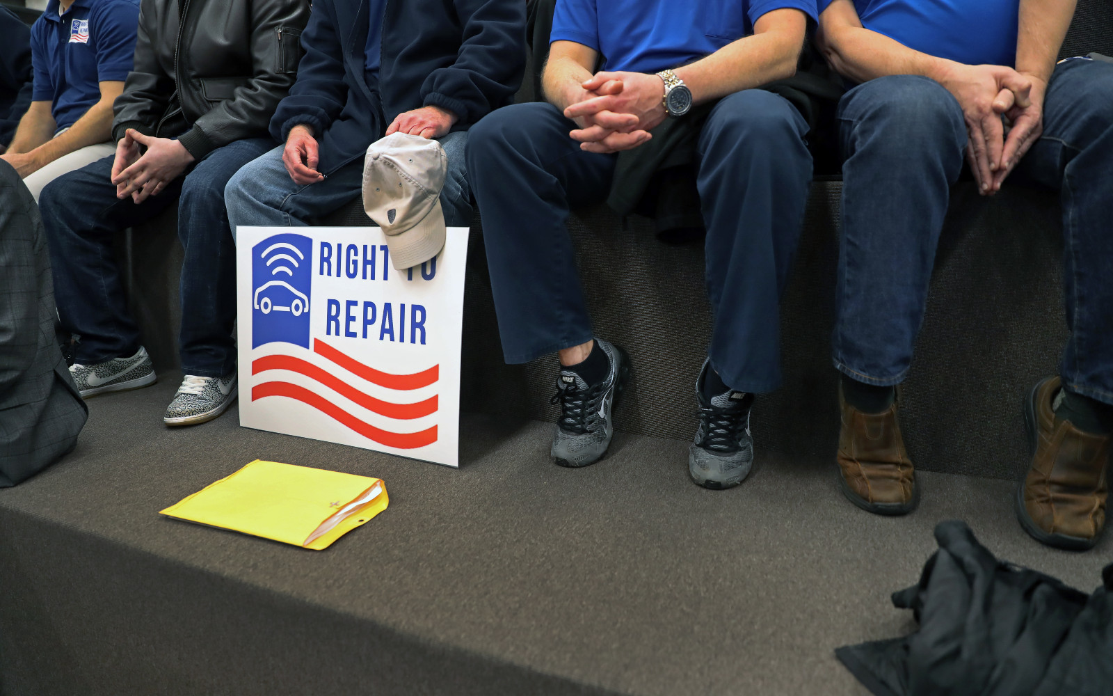 Torsos and legs of five men seated on a bench side by side, with a poster that says "Right to Repair" in front of one of them