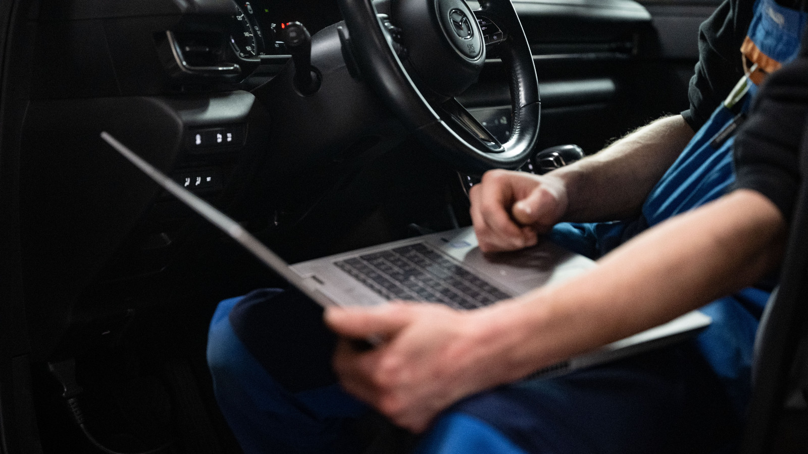The arms of a white man sitting in the driver's seat of a car holding a laptop