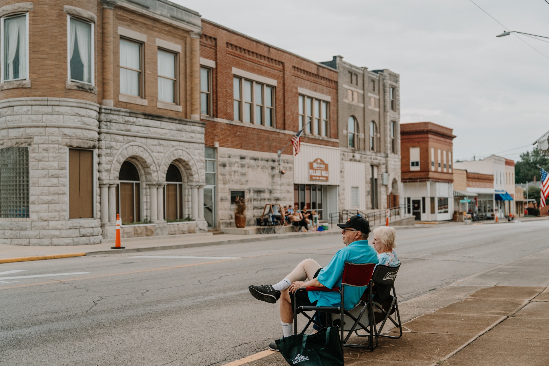An older couple sits in folding chairs on one side of an empty street looking across at people in chairs on the other side