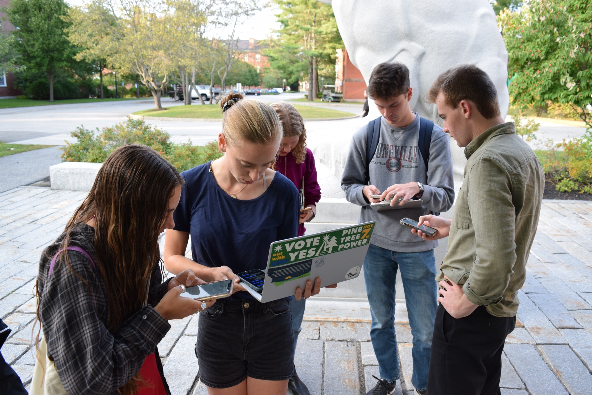 A group of young people stand together on a stone sidewalk holding phones and laptops. On the laptop is a large green sticker reading 'Vote Yes, Pine Tree Power'