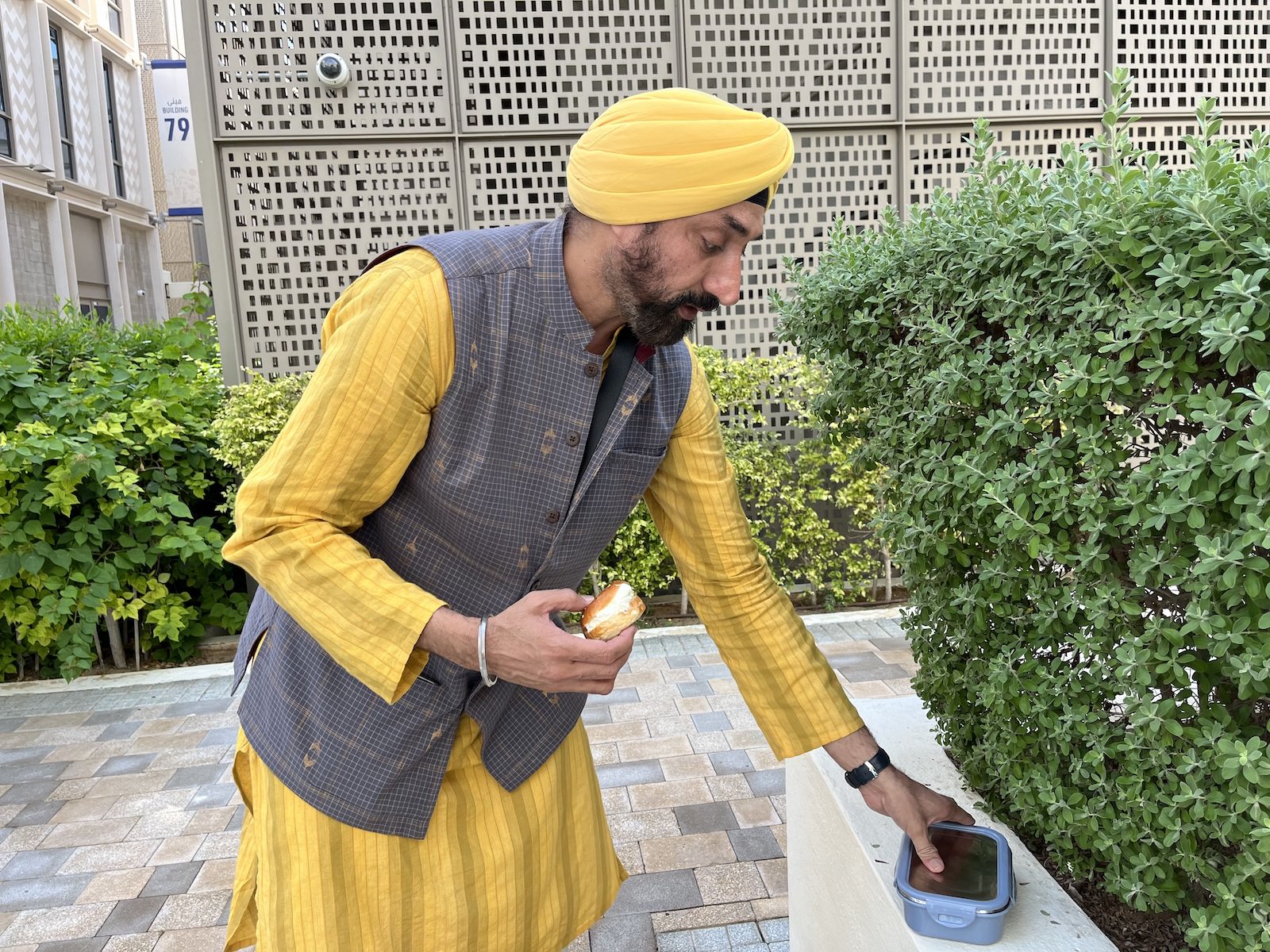 A man in a yellow turban and shirt reaches for a container in a courtyard with greenery and bricks